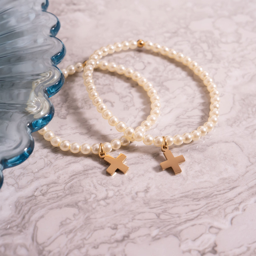 Two pearl bracelets with gold cross charms on a textured surface
