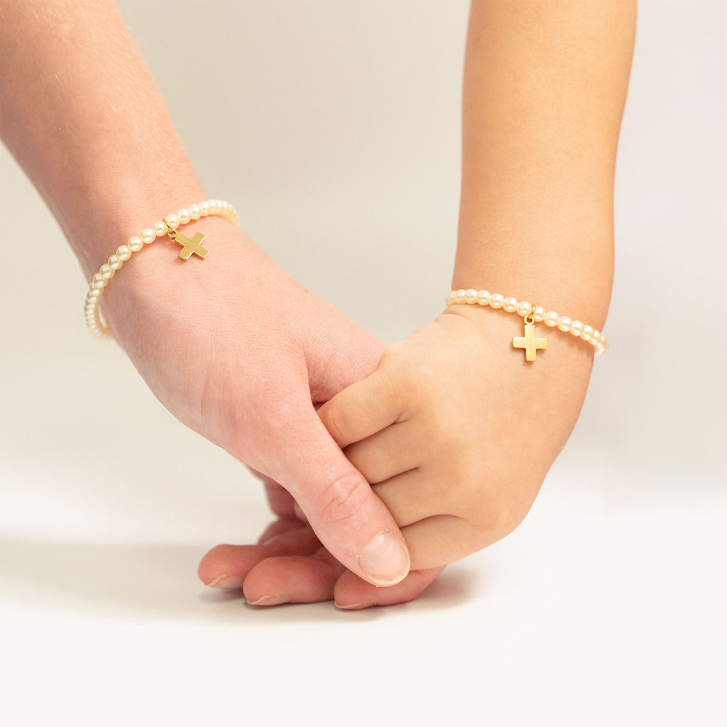 Two hands holding each other wearing pearl bracelets with cross charms on a light background