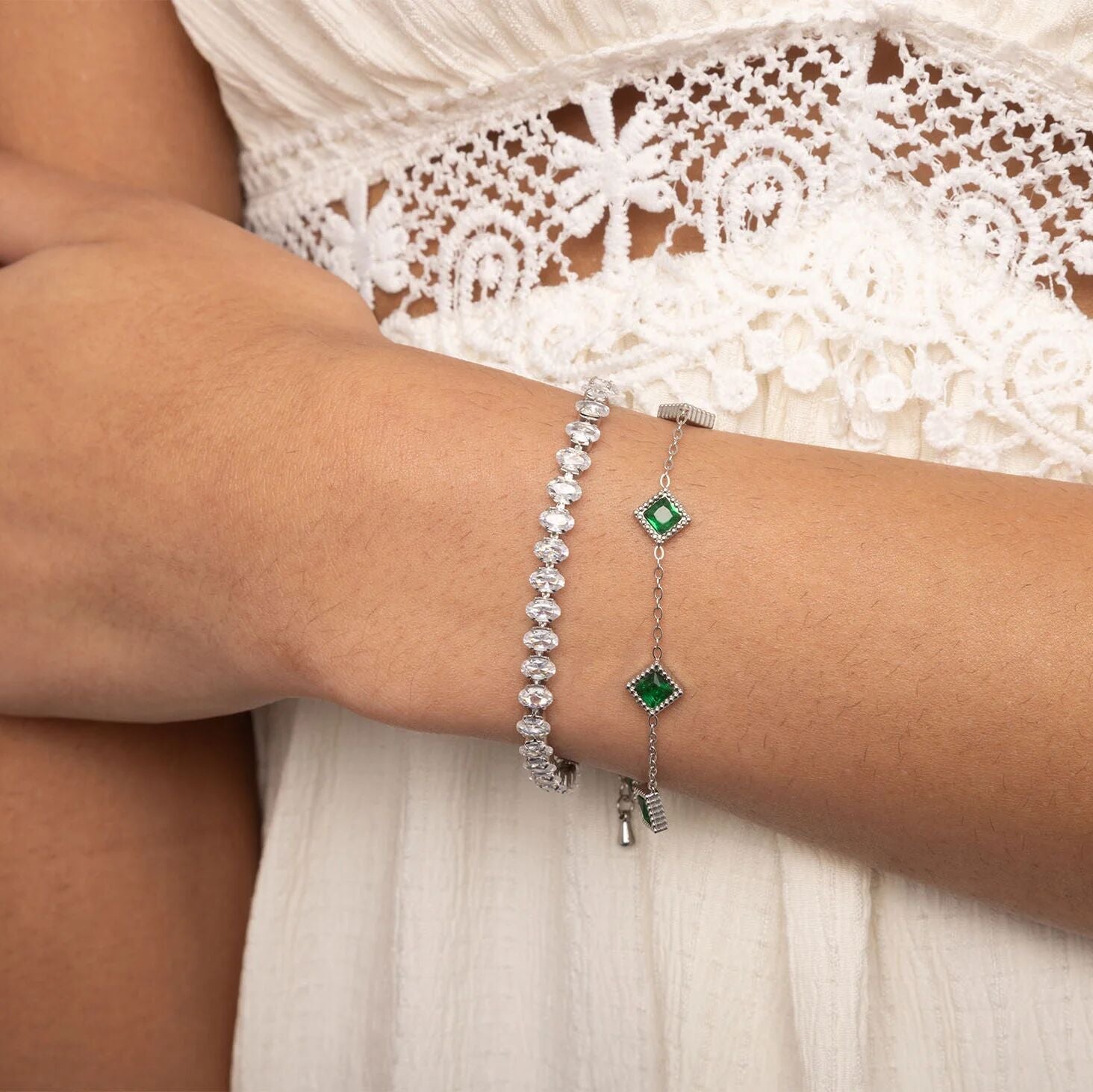 Close-up of a wrist wearing two silver bracelets with green gemstones, against a white lace garment.