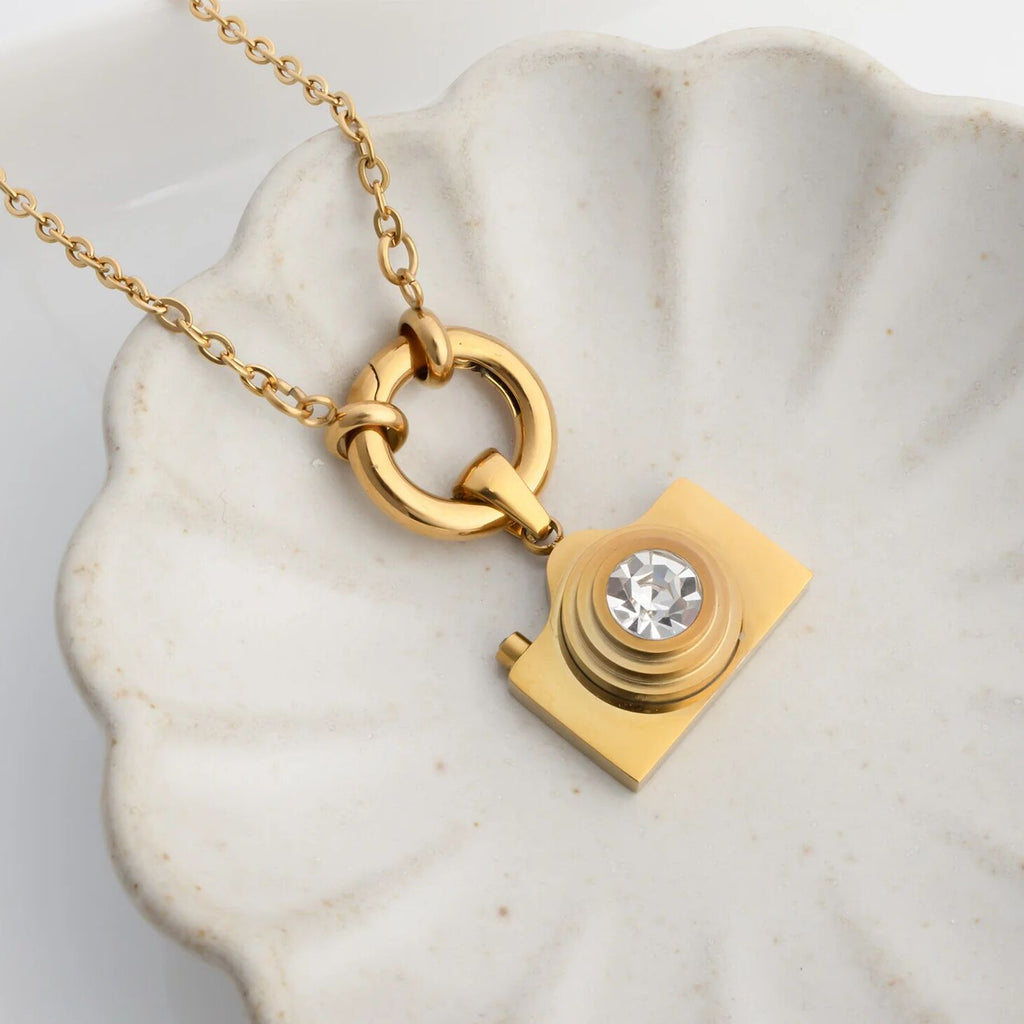 Gold charm holder necklace holding a gold camera-shaped pendant with a diamond on a marble surface