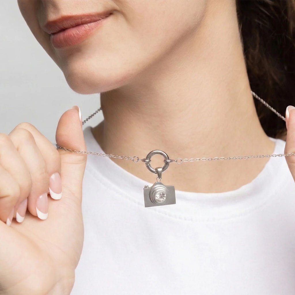 Person wearing a silver charm holder necklace with a camera-shaped pendant on a plain background