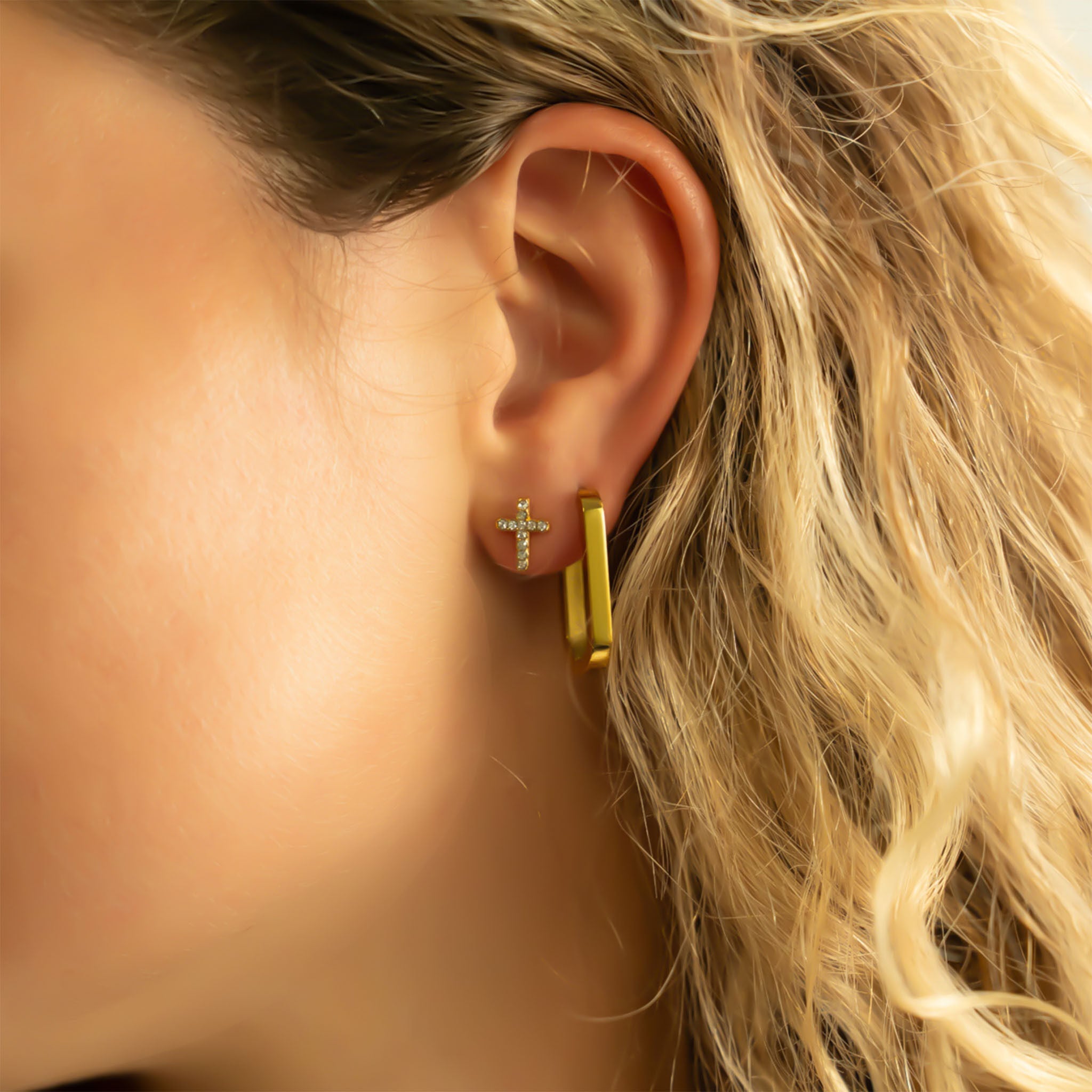 Close-up of a person wearing gold cross stud and hoop earrings with a blurred background