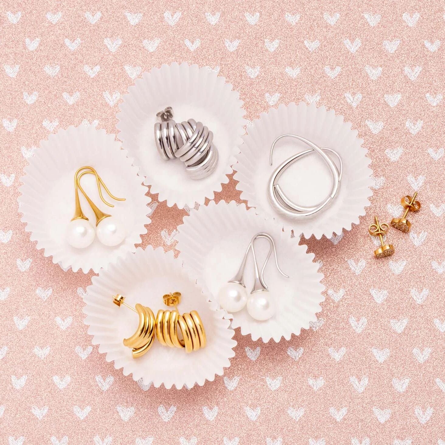 Gold and silver earrings on white paper cups against a pink background with heart patterns