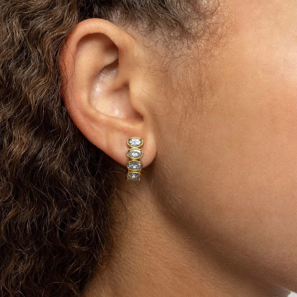 Close-up of an ear wearing gold Oval Stone Huggie Earrings with clear stones.