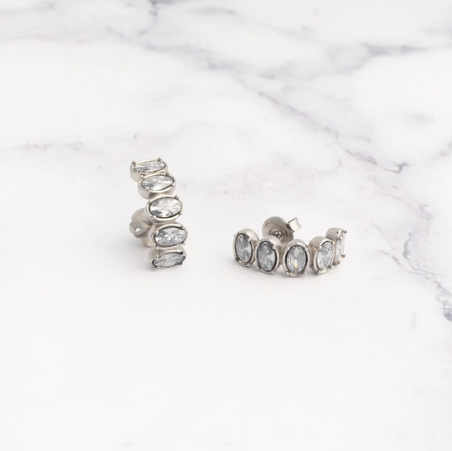 Pair of silver Oval Stone Huggie Earrings with clear stones on a marble surface