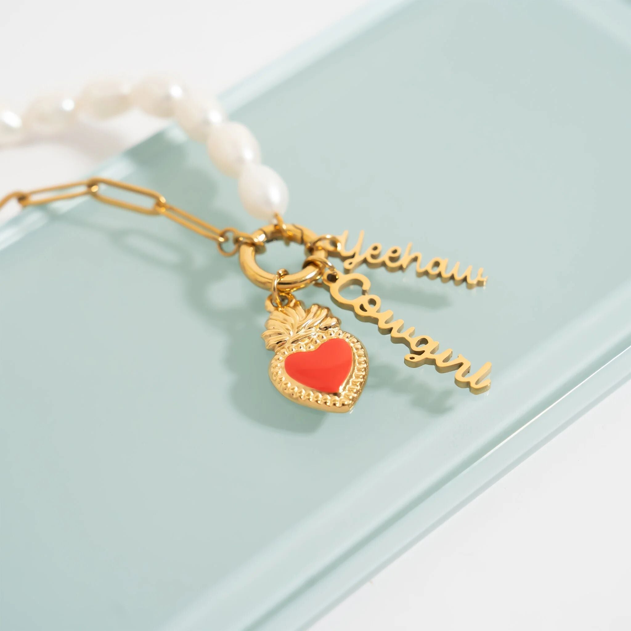 Gold charm with red heart and 'Yeehaw Cowgirl' text on a pearl necklace against a light blue background