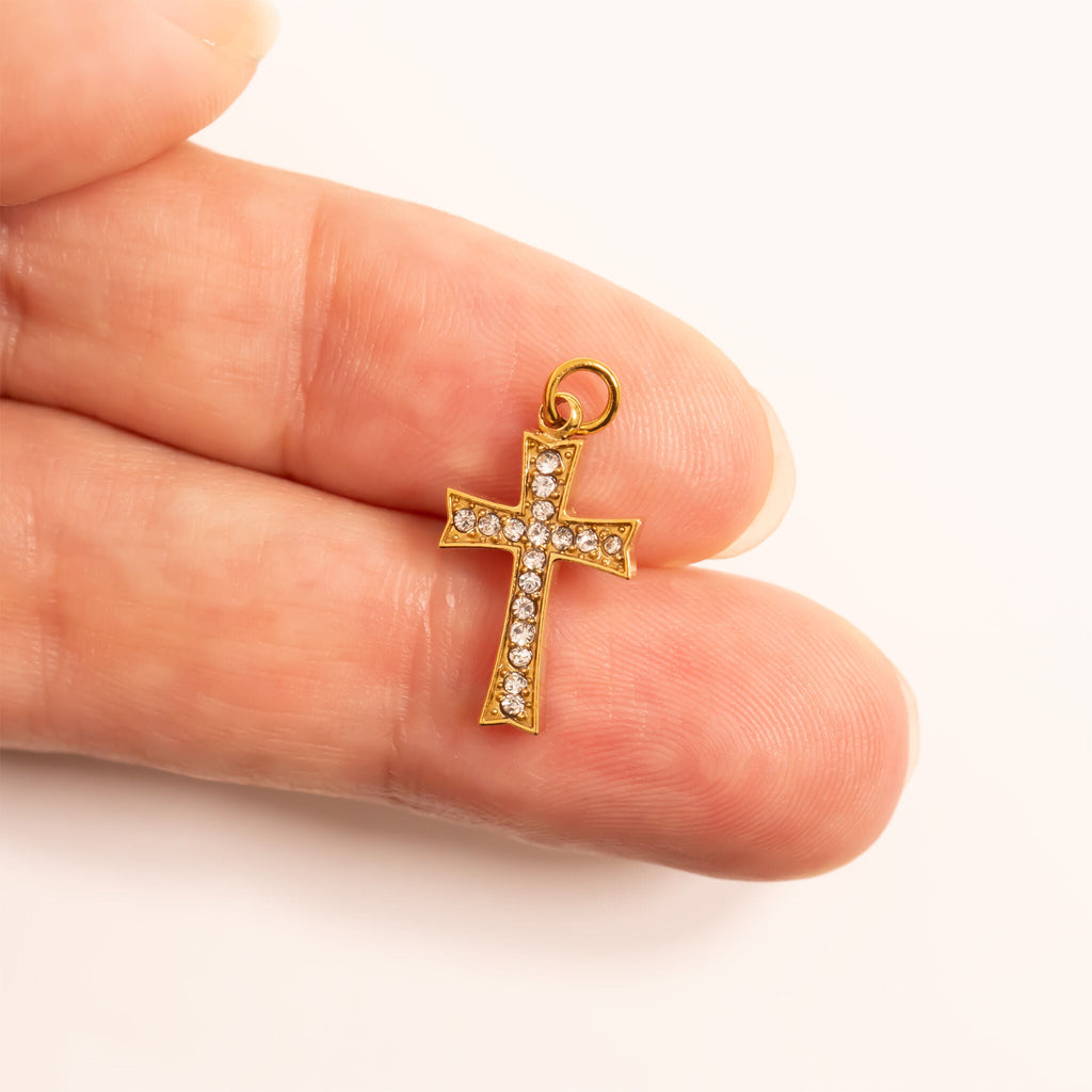 Gold cross pendant with clear stones held between two fingers for scale on a white background