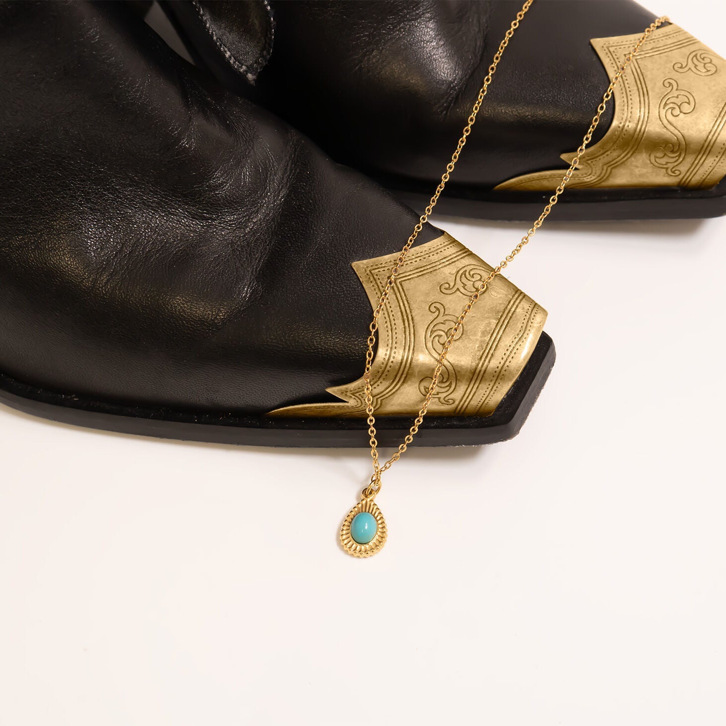 Gold necklace with a turquoise pendant draped over black leather cowboy boots on a white background