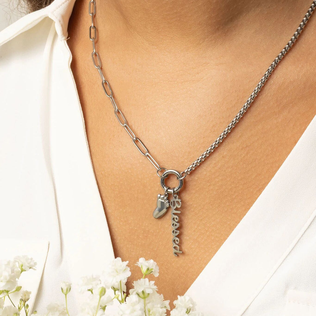 Silver charm holder necklace with a charms featuring a baby foot and the word 'blessed' worn by a person, with white flowers in the foreground.