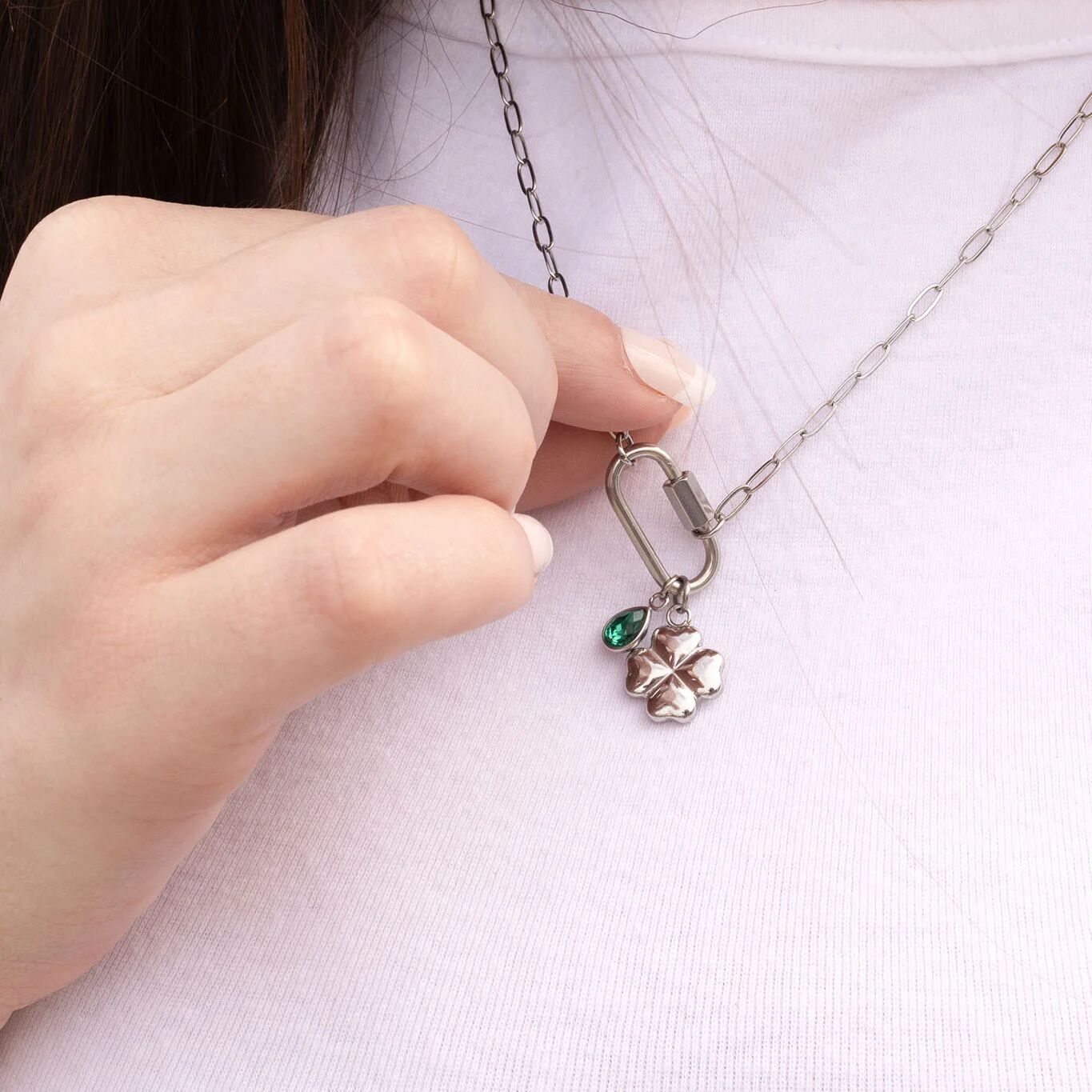 Hand holding a silver necklace with a clover pendant and green gemstone against a white background