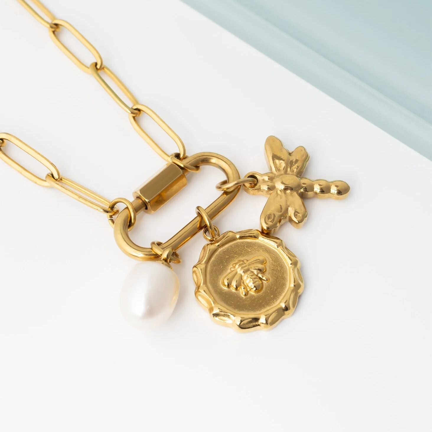 Gold necklace with a pearl, round bee, and dragonfly charm on a light background