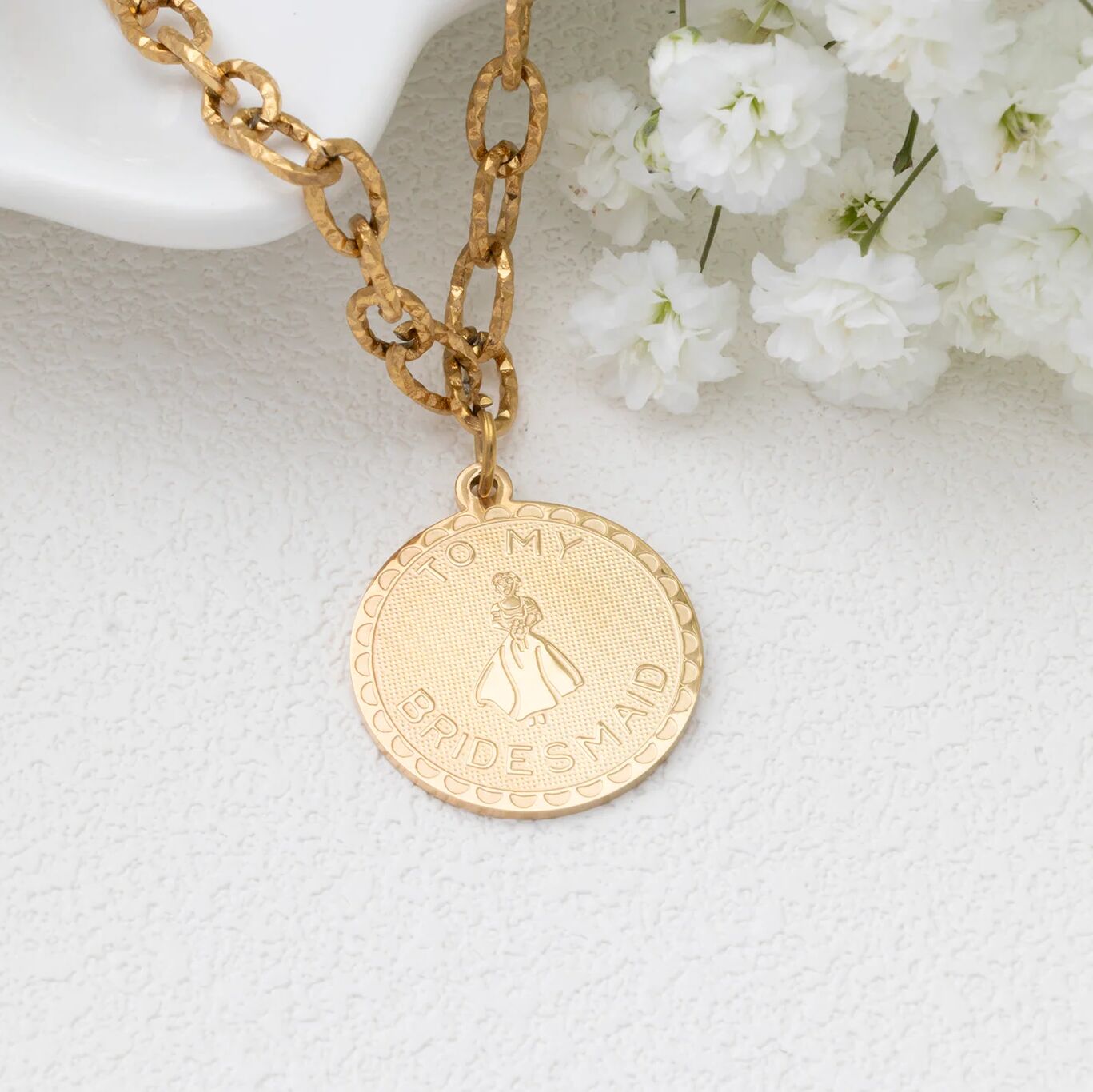 Gold necklace with a 'To My Bridesmaid' pendant on a white textured surface with white flowers.