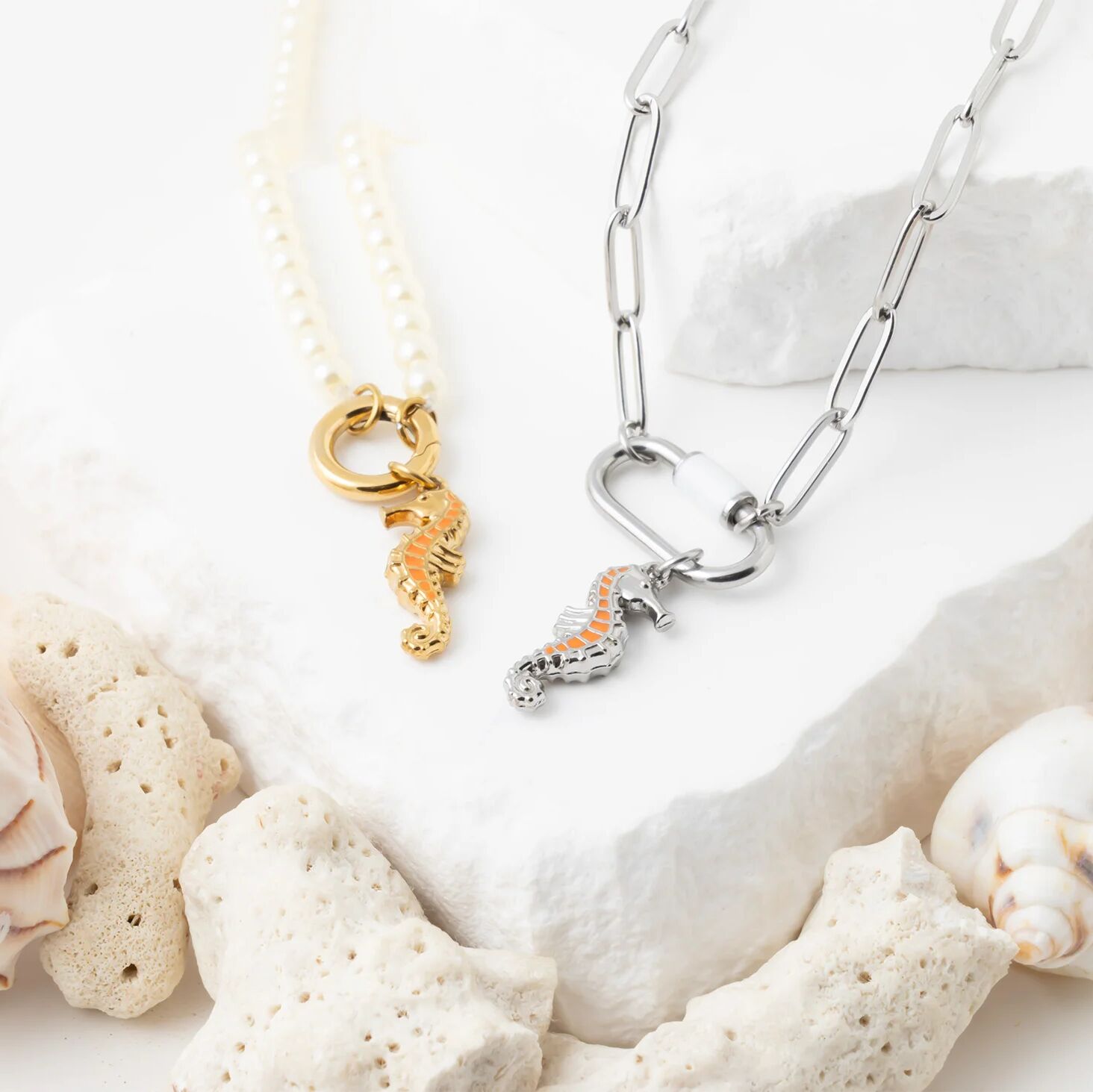 Two seahorse-themed necklaces on a white surface with shells and sand.