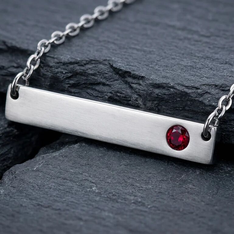 Silver necklace with a horizontal bar pendant featuring a red gemstone on a dark stone surface.